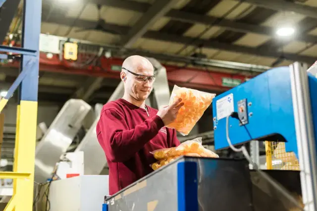 A worker in safety glasses inspects bags of orange snack chips at a factory, surrounded by industrial equipment.