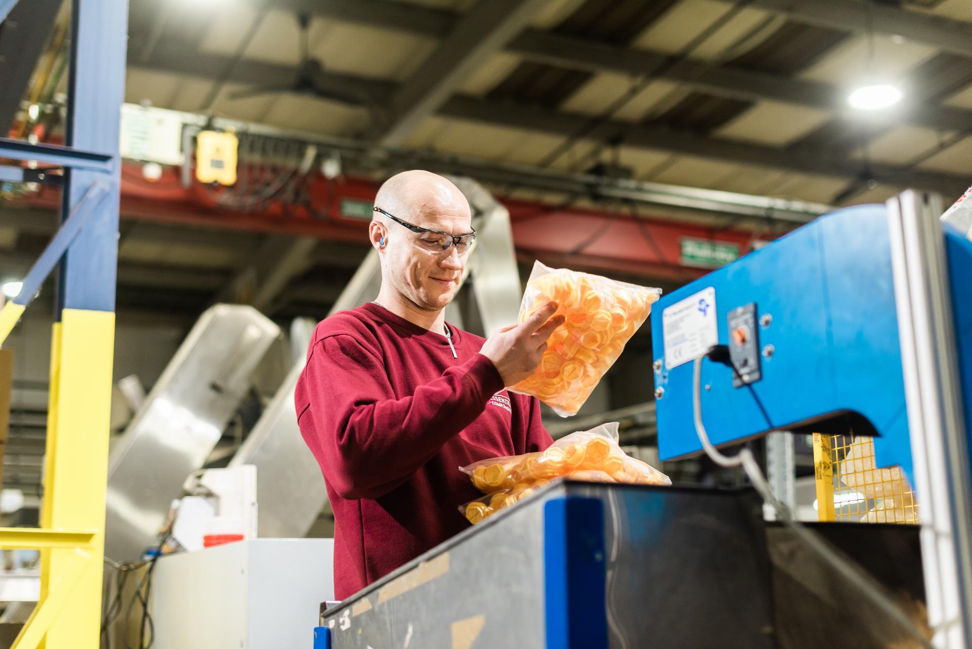 A person wearing safety goggles examines packaged products in a factory, standing near blue industrial equipment.