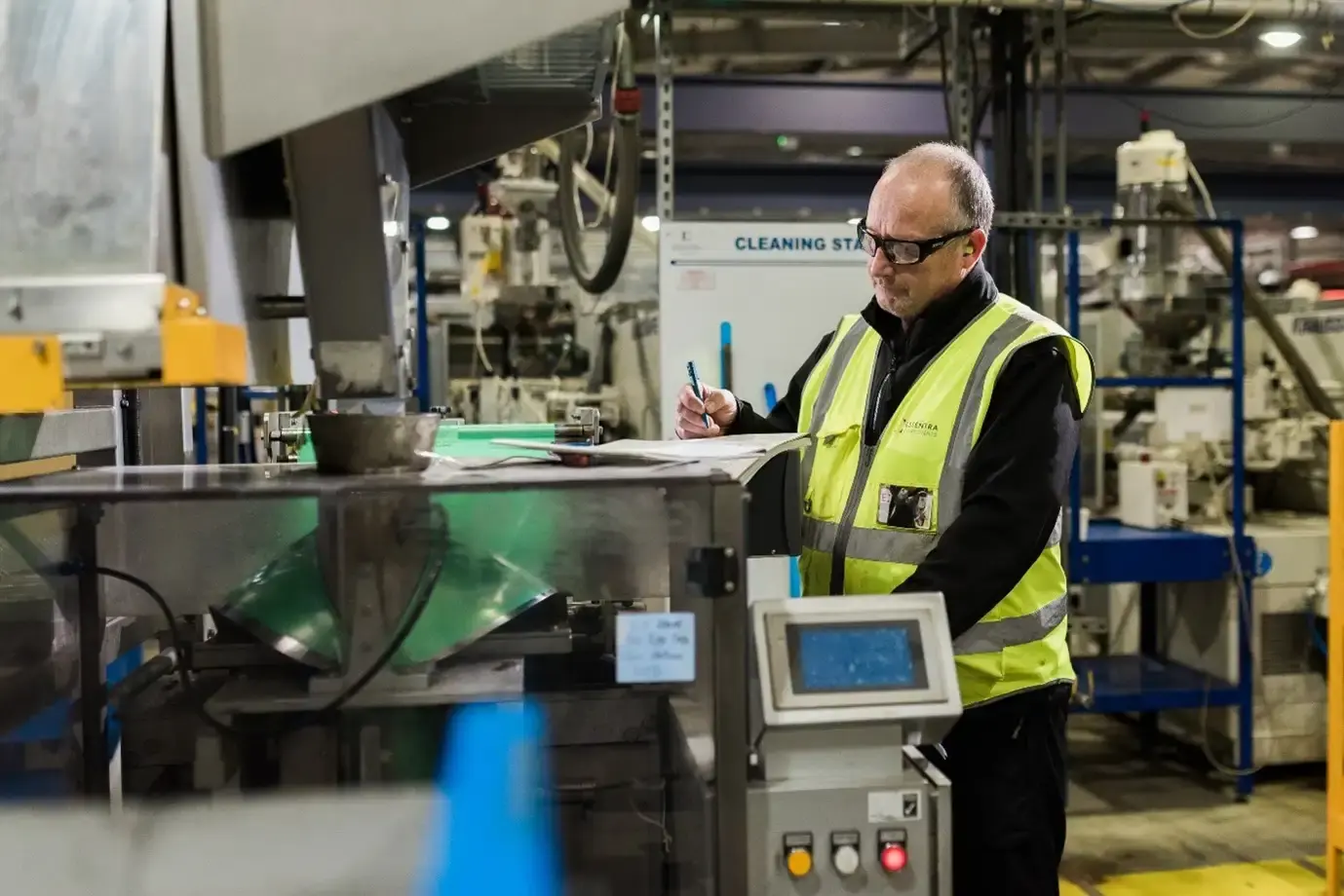 A worker in a reflective vest and safety glasses writes on a clipboard in an industrial setting with machinery around.